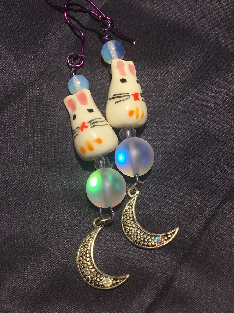 photo of hand made earrings. vertical bunny charm sitting on a moonstone with a moon charm at the end to dangle. the wire is purple.